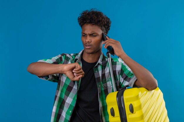 Młody człowiek afroamerykański podróżnik stojący z walizką patrząc na ukrytą dłoń, przypominający sobie o czasie podczas rozmowy przez telefon komórkowy na niebieskim tle