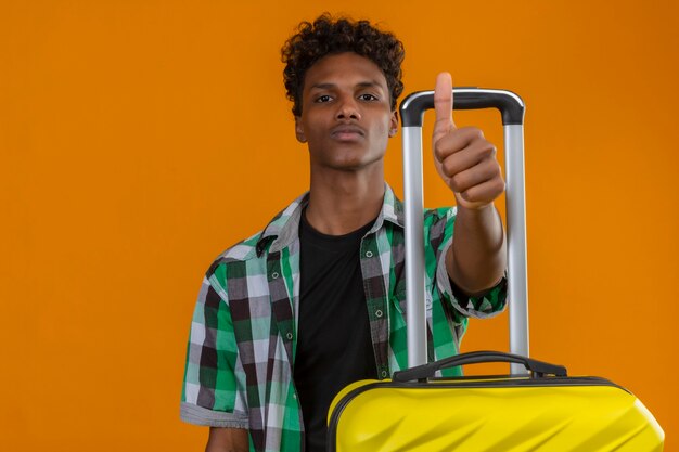 Młody człowiek afroamerykański podróżnik z walizką z pewną poważną miną na twarzy pokazując kciuk do góry