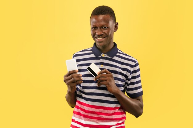 Młody człowiek afroamerykański z smartphone i debet karty na białym tle na żółtym tle studio