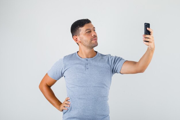 Młody człowiek biorąc selfie z ręką na talii w szarej koszulce.