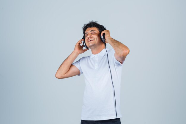 Młody człowiek ciesząc się muzyką ze słuchawkami w białej koszulce i patrząc szczęśliwy, widok z przodu.