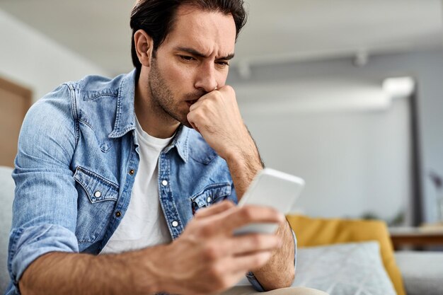 Młody człowiek czuje się zmartwiony podczas czytania wiadomości tekstowej na telefonie komórkowym w domu