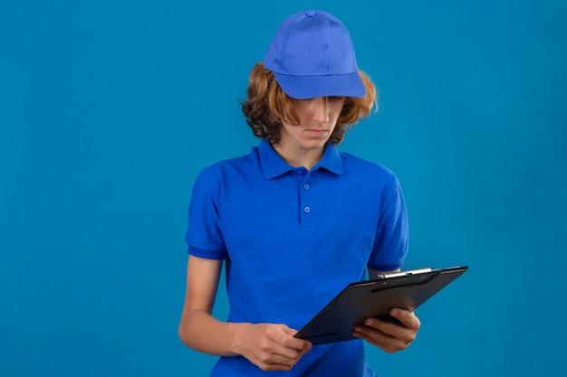 Młody człowiek dostawy na sobie niebieską koszulkę polo i czapkę patrząc na schowek w ręku z niezadowoloną twarzą stojącą na na białym tle niebieskim tle