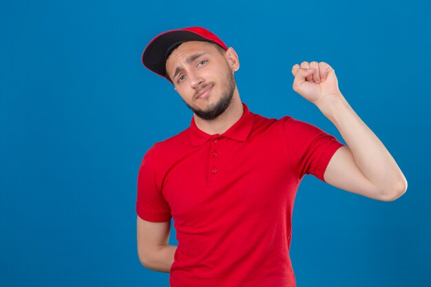 Młody człowiek dostawy ubrany w czerwoną koszulkę polo i czapkę podnosząc pięść po koncepcji zwycięzcy zwycięstwa na na białym tle niebieskim tle