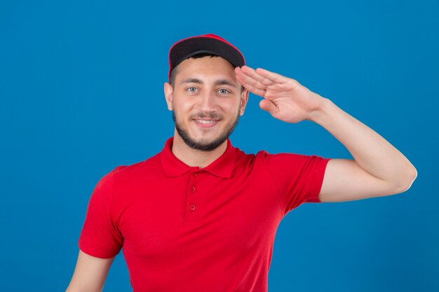 Młody człowiek dostawy ubrany w czerwoną koszulkę polo i czapkę salutowania patrząc pewnie na na białym tle niebieskim tle