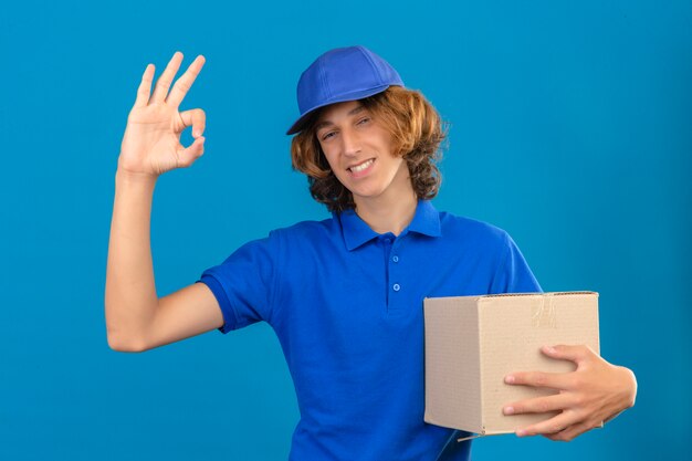 Młody człowiek dostawy ubrany w niebieską koszulkę polo i czapkę trzymając karton robi ok znak uśmiechnięty przyjazny na pojedyncze niebieskie tło