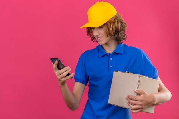 Bezpłatne zdjęcie młody człowiek dostawy ubrany w niebieską koszulkę polo i żółtą czapkę trzymający karton patrząc na ekran swojego smartfona z uśmiechem na twarzy stojącej na izolowanym różowym tle