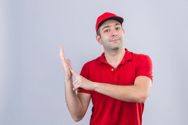 Młody człowiek dostawy w czerwonej koszulce polo i czapce, wskazując palcem na dłoń, patrząc pewnie na na białym tle
