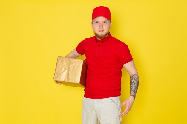 młody człowiek dostawy w czerwonej polo czerwonej czapce białe dżinsy trzyma pudełko na żółtym