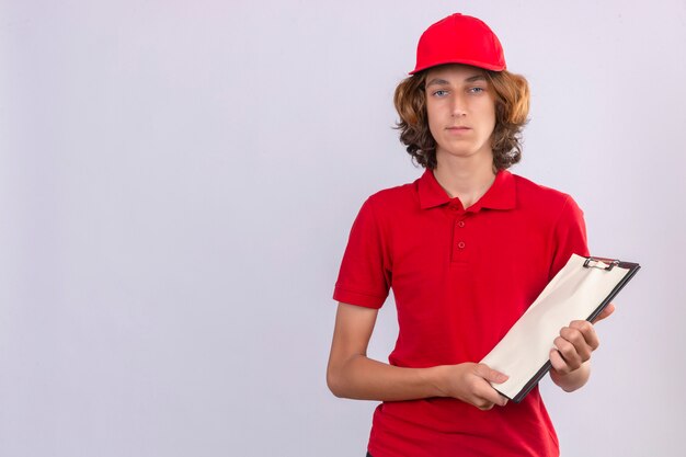 Młody człowiek dostawy w czerwonym mundurze gospodarstwa schowka patrząc na kamery sceptyczny i zdenerwowany na na białym tle