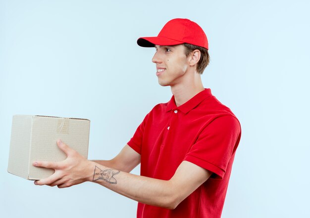 Młody człowiek dostawy w czerwonym mundurze i czapce dając karton klientowi uśmiechając się stojąc na białej ścianie