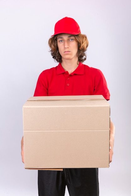 Młody człowiek dostawy w czerwonym mundurze, posiadający duży karton patrząc na kamery ze smutnym wyrazem twarzy stojącej na białym tle