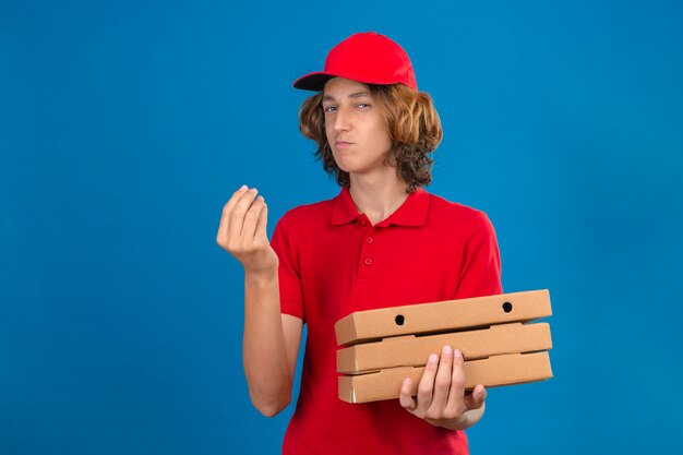 Młody człowiek dostawy w czerwonym mundurze, trzymając pudełka po pizzy robi pyszne gesty ręką uśmiechając się na na białym tle niebieskim tle