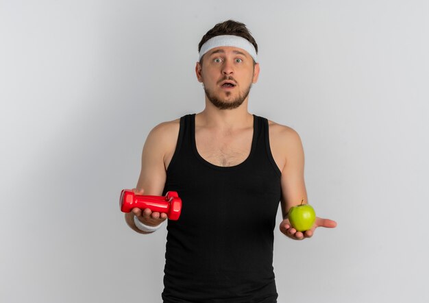 Młody człowiek fitness z pałąkiem na głowę trzymając zielone jabłko i hantle patrząc na kamery zdezorientowany stojąc na białym tle
