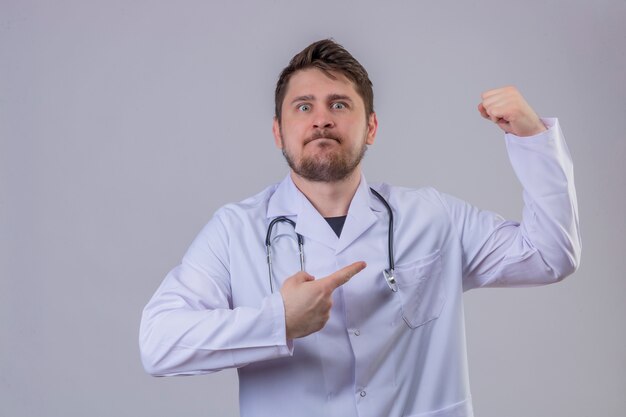 Młody człowiek lekarz ubrany w biały fartuch i stetoskop wskazując palcem na bicepsy, koncepcja zwycięzcy