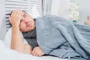Bezpłatne zdjęcie młody człowiek leżący na łóżku z bólem głowy i gorączką dotykając jego czoło