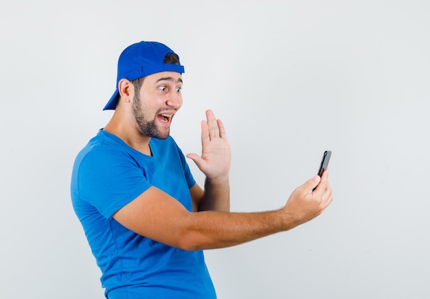 Młody człowiek macha ręką na czacie wideo w niebieskiej koszulce i czapce i wygląda wesoło