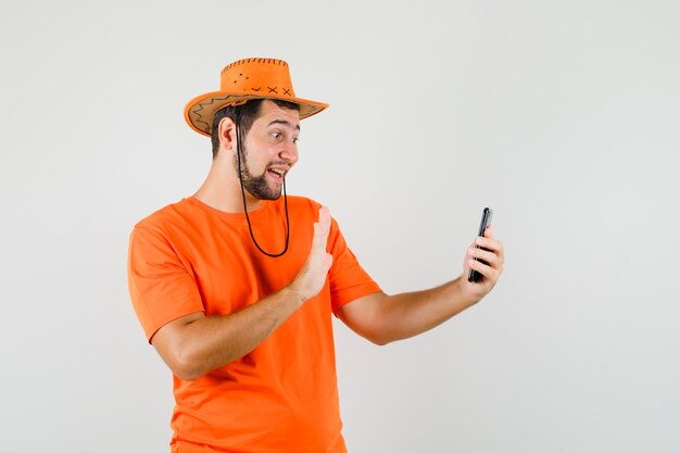 Młody człowiek macha ręką podczas robienia selfie w pomarańczowej koszulce, kapeluszu i wygląda wesoło