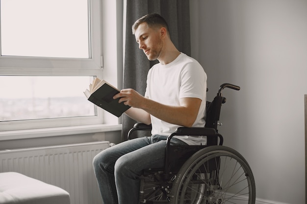 Młody człowiek niepełnosprawny. Mężczyzna czyta książkę na wózku inwalidzkim, przebywa w domu.