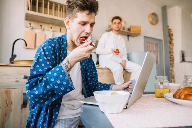Młody człowiek patrzeje jego przyjaciela je sałatki podczas gdy pracujący na laptopie