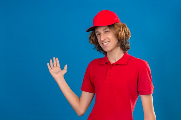 Młody człowiek pewny dostawy w czerwonym mundurze, uśmiechnięty przyjazny prezentując coś ręką na odosobnionej niebieskiej ścianie