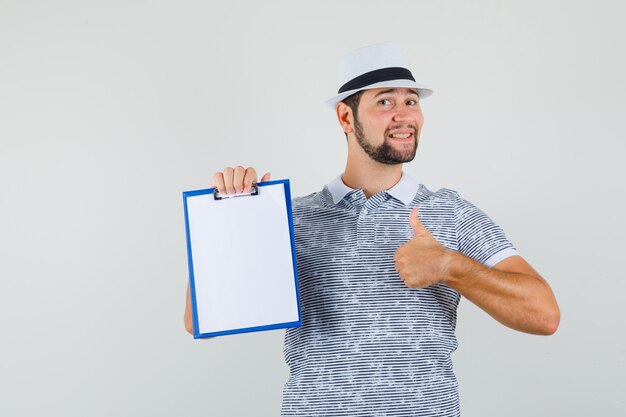 Młody człowiek pokazując kciuk do góry trzymając notebook w t-shirt, kapelusz i patrząc pewnie, widok z przodu.