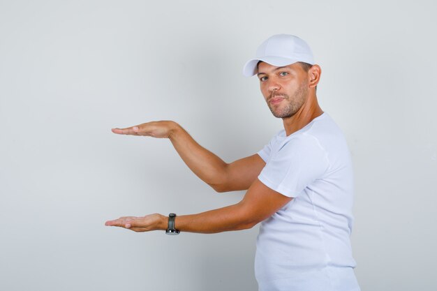 Młody człowiek pokazujący znak rozmiaru z rękami w białej koszulce, czapkę, widok z przodu.