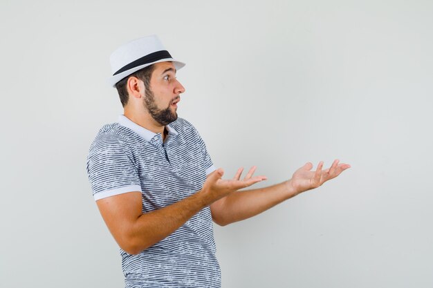 Młody człowiek pokazuje bezradny gest w t-shirt, kapelusz i patrząc zdezorientowany. przedni widok.