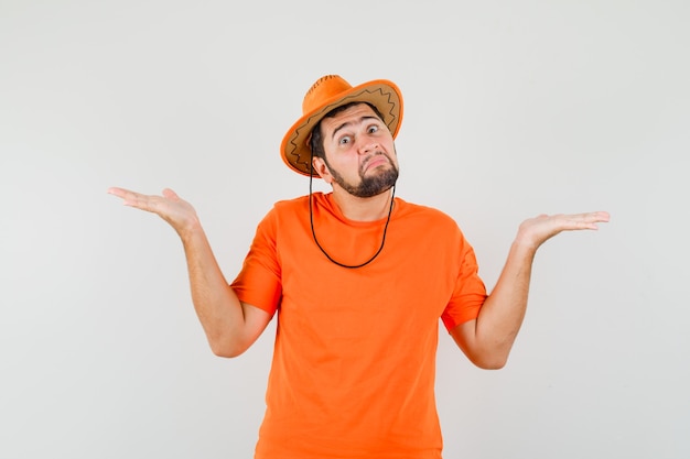 Młody człowiek pokazuje bezradny gest wzruszając ramionami w pomarańczowej koszulce, kapeluszu i patrząc zdezorientowany, widok z przodu.