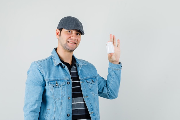 Młody człowiek pokazuje butelkę tabletek w kurtce, czapkę i zadowolony, widok z przodu.