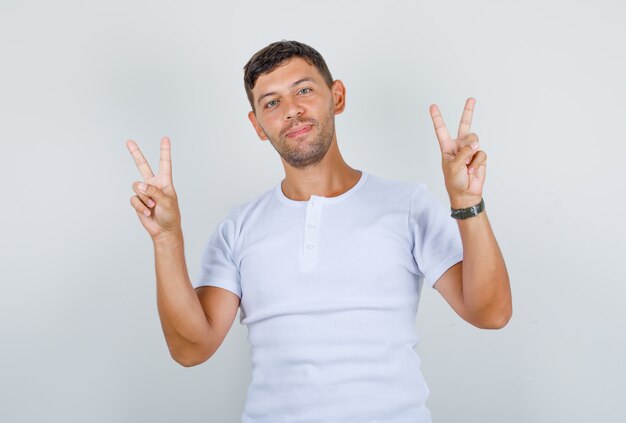 Młody człowiek pokazuje palce robi znak zwycięstwa w białej koszulce i wygląda szczęśliwy, widok z przodu