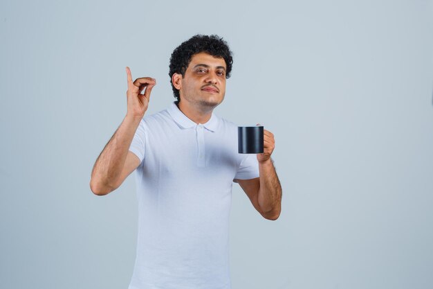 Młody człowiek pokazuje pyszny gest i trzyma filiżankę herbaty w białej koszulce i dżinsach i wygląda na szczęśliwego, widok z przodu.