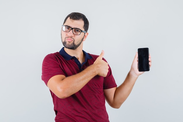 Młody człowiek pokazuje telefon, podczas gdy kciuk w górę w czerwonej koszulce, optyczne okulary i wygląda zadowolony. przedni widok.