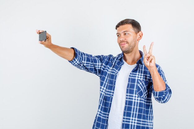 Młody człowiek pokazuje znak v podczas robienia selfie w koszuli i wygląda wesoło. przedni widok.