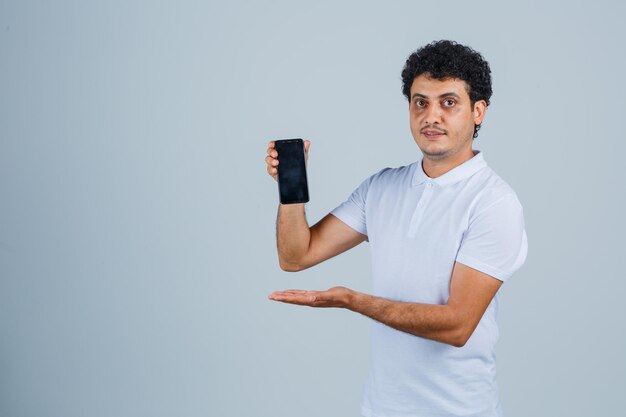 Młody człowiek prezentuje telefon komórkowy w białej koszulce i wygląda pewnie. przedni widok.