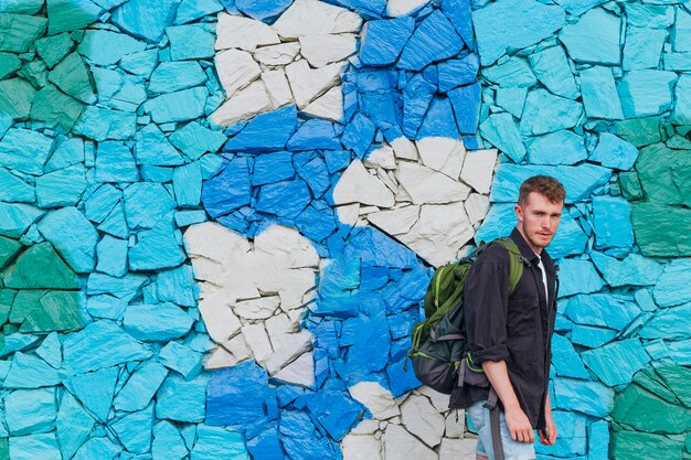 Młody człowiek stoi blisko malującej kamiennej ściany z podróż plecakiem
