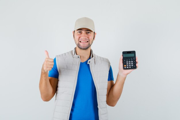 Młody człowiek trzyma kalkulator, pokazując kciuk w t-shirt, kurtkę i wesoły wyglądający. przedni widok.
