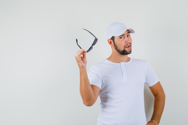 Młody człowiek trzyma okulary podczas pozowanie w t-shirt, czapkę i wyglądający pewnie. przedni widok. miejsce na tekst