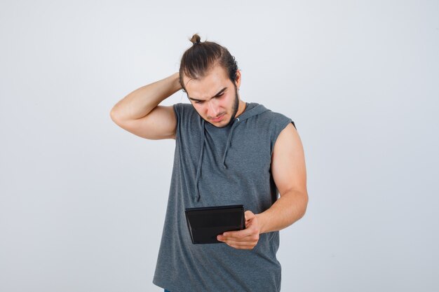 Młody człowiek trzyma rękę za głową, patrząc na kalkulator w bluzie z kapturem i patrząc zamyślony, widok z przodu.