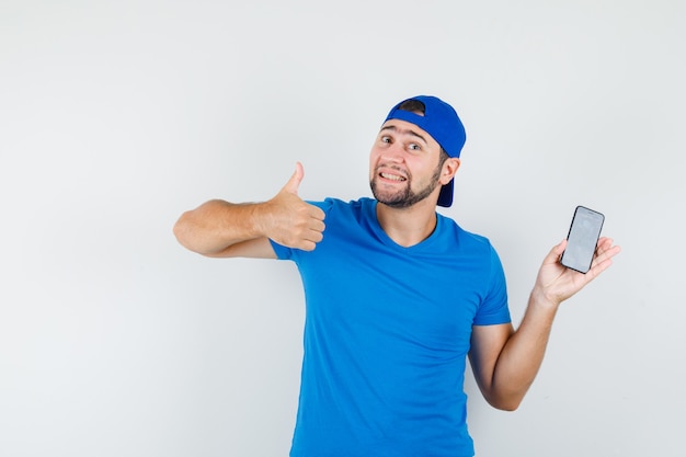 Młody człowiek trzyma telefon komórkowy z kciukiem w niebieską koszulkę i czapkę i wygląda zadowolony