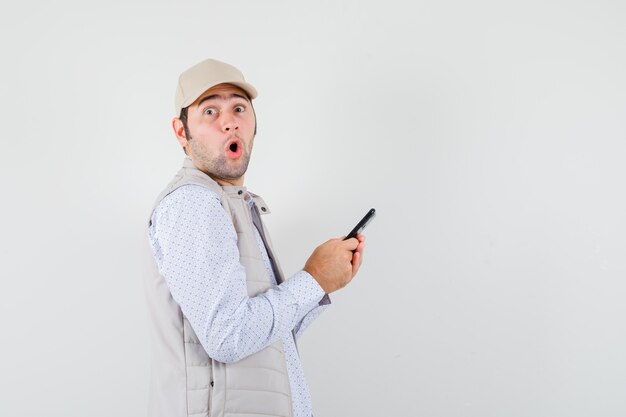 Młody człowiek, trzymając telefon komórkowy pod ręką w beżowej kurtce i czapce i patrząc zaskoczony. przedni widok.