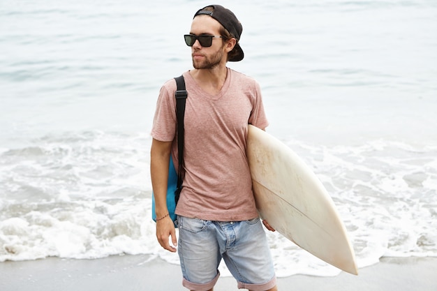 Młody człowiek ubrany w modne okulary przeciwsłoneczne i snapback, trzymając w ręku deskę surfingową