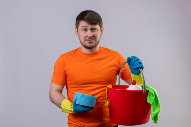 Młody człowiek ubrany w pomarańczową koszulkę i gumowe rękawiczki trzymający wiadro z narzędziami do czyszczenia i gąbką niezadowolony i bardzo niespokojny stojący nad białą ścianą