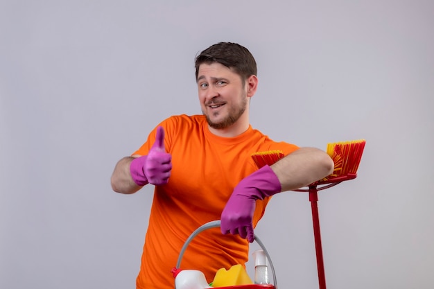 Młody człowiek ubrany w pomarańczową koszulkę i gumowe rękawiczki trzymający wiadro z narzędziami do czyszczenia i mopem pozytywny i szczęśliwy, uśmiechnięty pokazując kciuki do góry stojąc nad białą ścianą