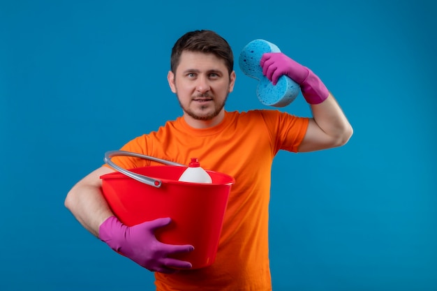 Młody człowiek ubrany w pomarańczowy t-shirt i gumowe rękawiczki, trzymając wiadro z narzędziami do czyszczenia i gąbką