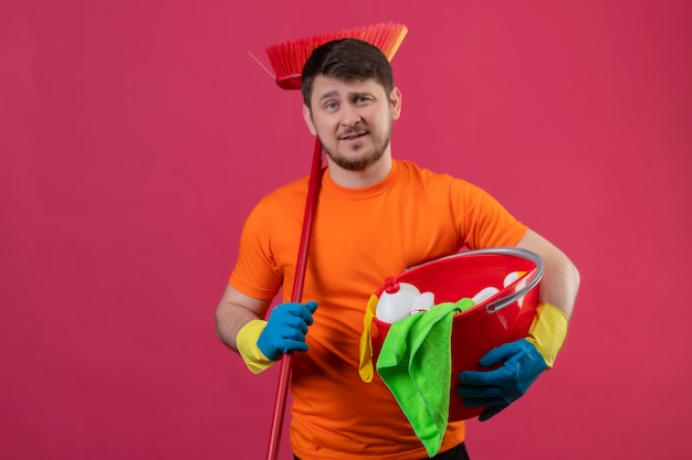 Młody człowiek ubrany w pomarańczowy t-shirt i gumowe rękawiczki, trzymając wiadro z narzędziami do czyszczenia i mopem