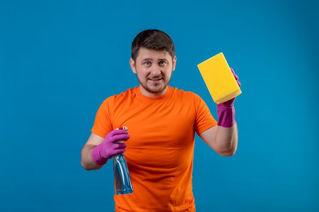 Młody człowiek ubrany w pomarańczowy t-shirt i rękawice gumowe, trzymając spray do czyszczenia i gąbkę