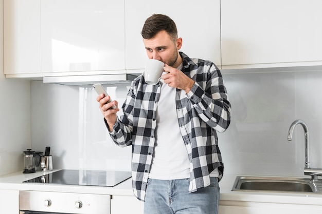 Młody człowiek używa telefon komórkowego podczas gdy pijący kawę w kuchni