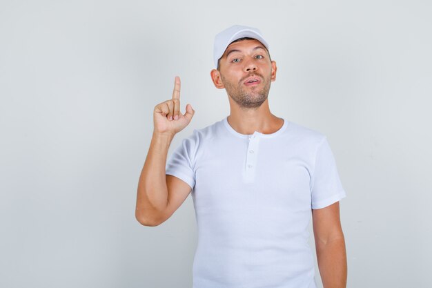 Młody człowiek w białej koszulce, czapka skierowana w górę palcem wskazującym i pewny, widok z przodu.
