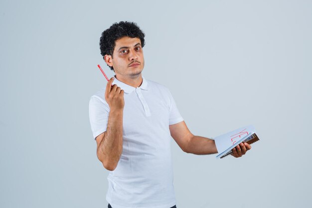 Młody człowiek w białej koszulce i dżinsach gryzie długopis, trzymając notatnik i myśląc o czymś i patrząc zamyślony, widok z przodu.